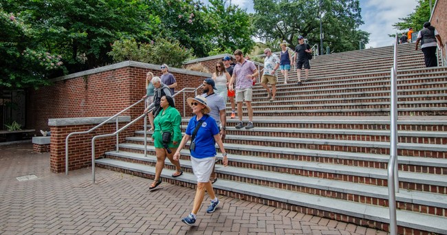 Tour guests and guide walking down brick steps with green trees and cloudy sky in the background.