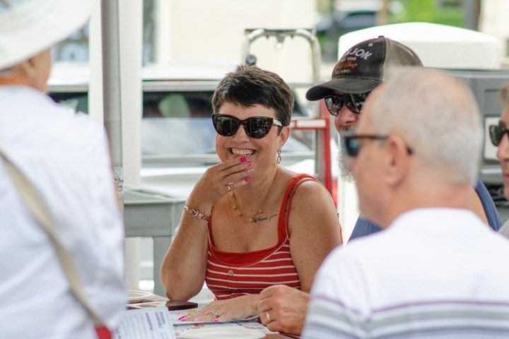 Group of people in sunglasses sitting around a table outdoors, with one woman smiling.