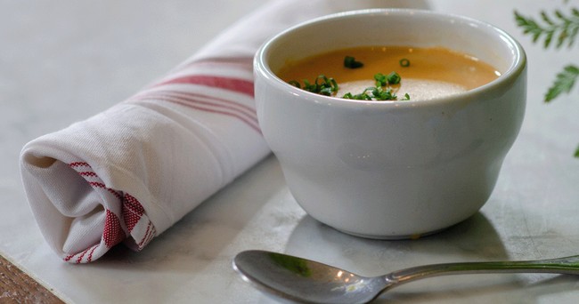Soup in a white cup with herbs, spoon, and a rolled napkin on a table.