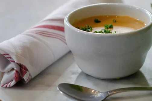 Soup in a white cup with herbs, spoon, and a rolled napkin on a table.