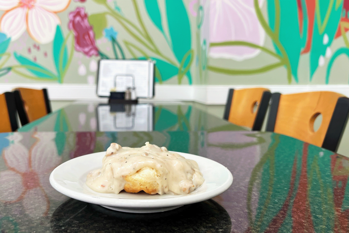 Biscuit with gravy on a plate, colorful floral mural and wooden chairs in the background.