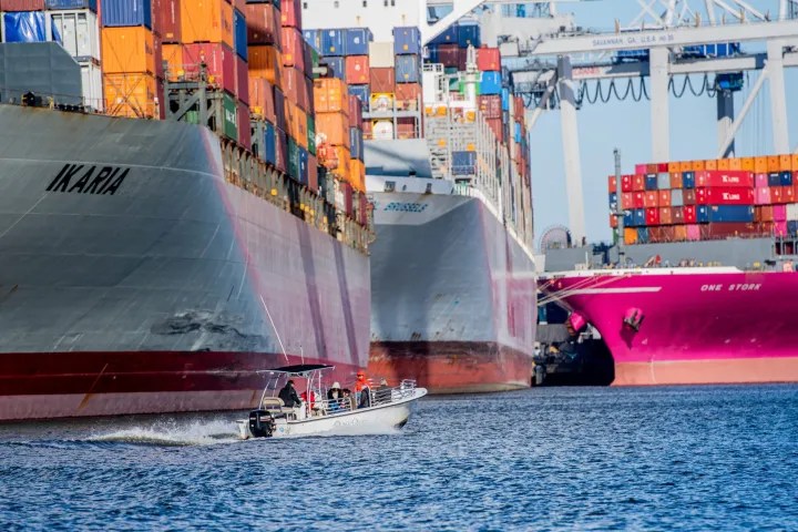 Container ships and a small boat in a busy port with stacked containers visible.
