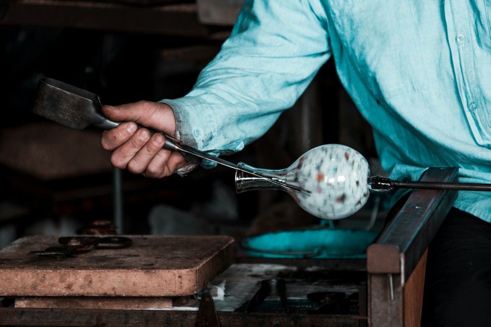 Person shaping glass with a metal tool and rod in a glass blowing workshop setting.