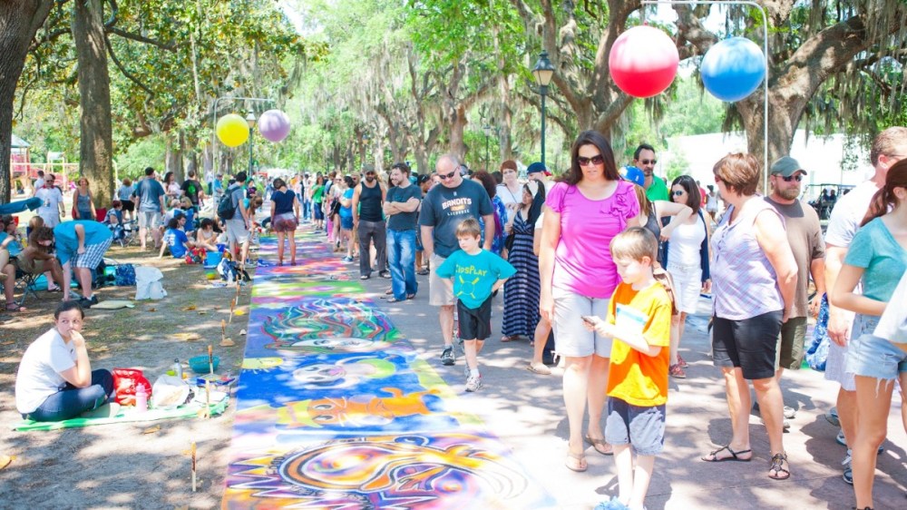 People walk along a path with colorful chalk art at an outdoor festival under trees.