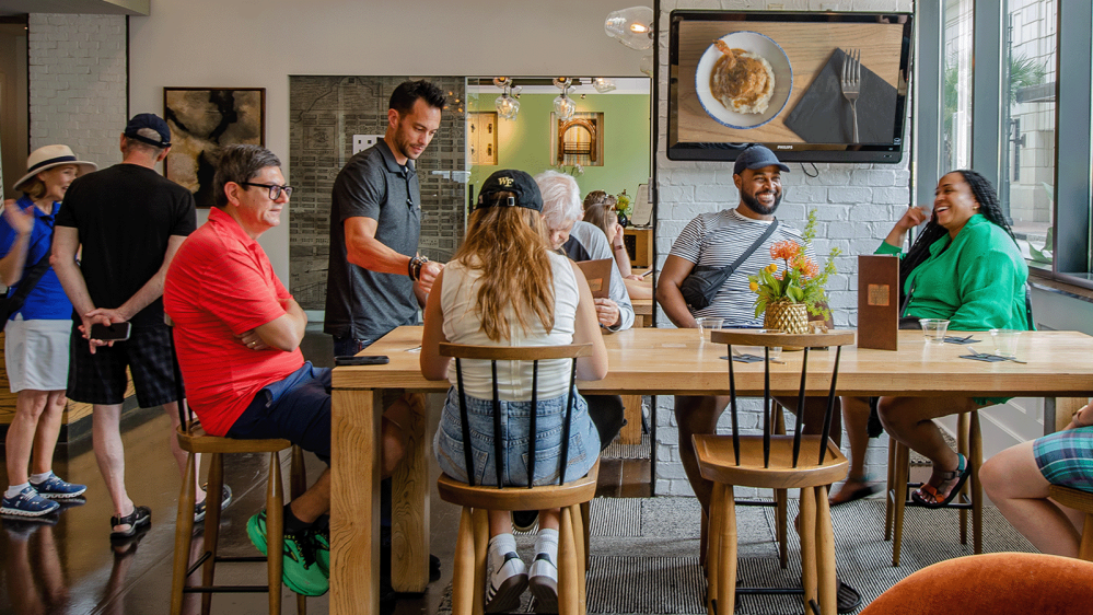 People sitting and standing around a table in a busy cafe, with a TV displaying food above.