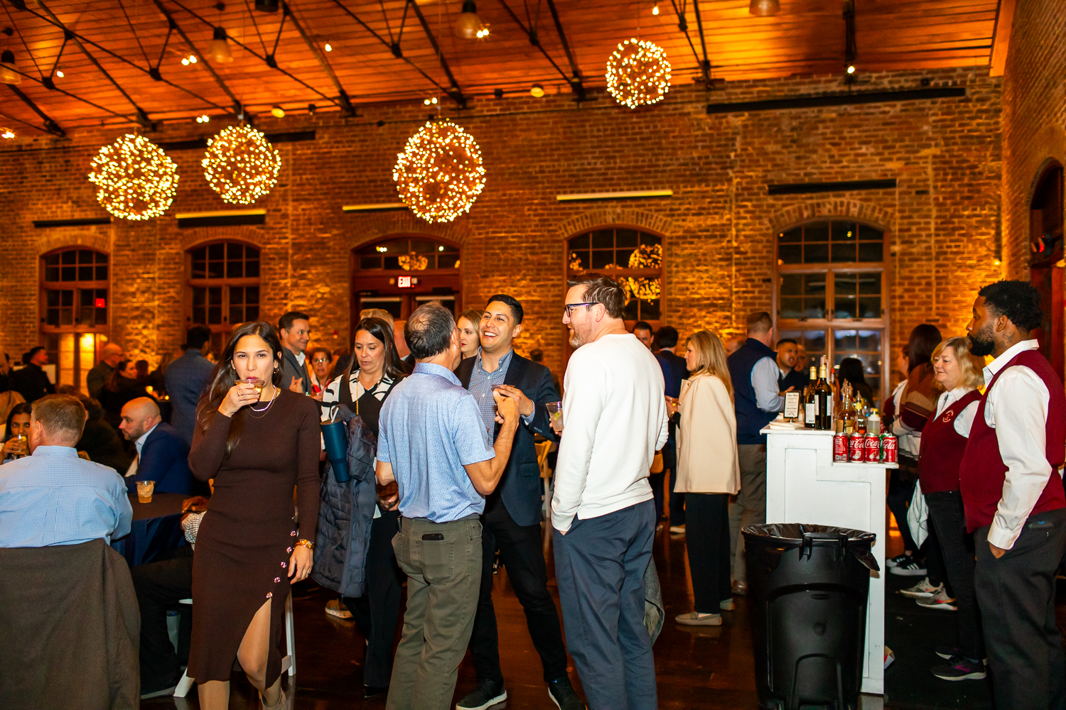 People socializing at an indoor event with drinks, brick walls, and warm lighting.