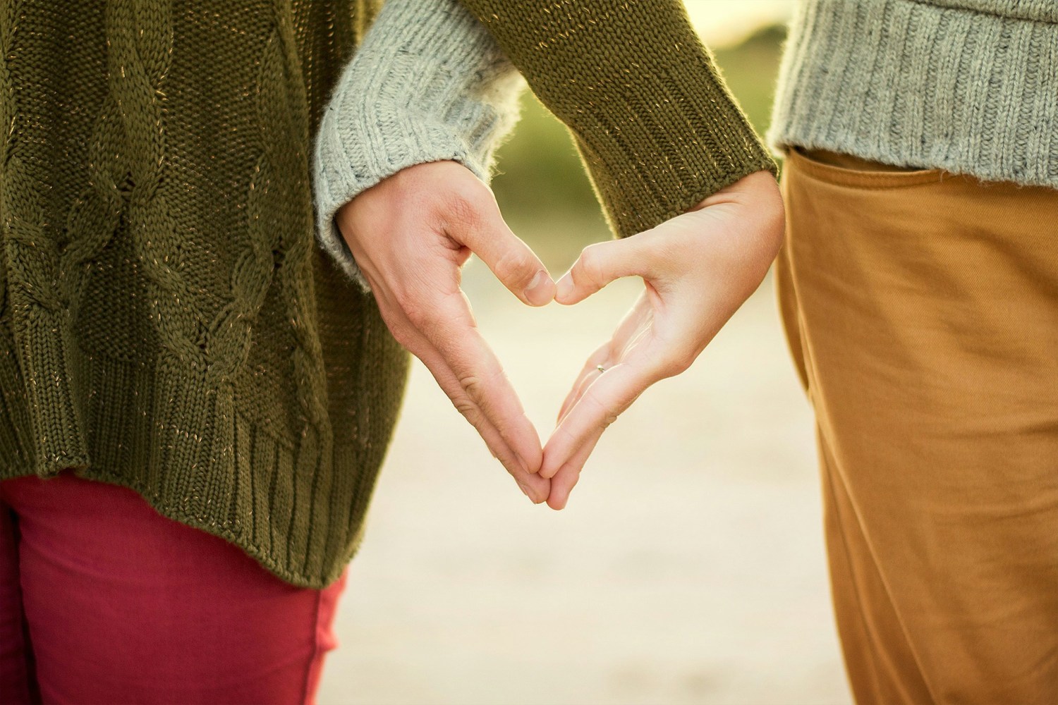 Two people forming a heart shape with hands, wearing sweaters and standing side by side.