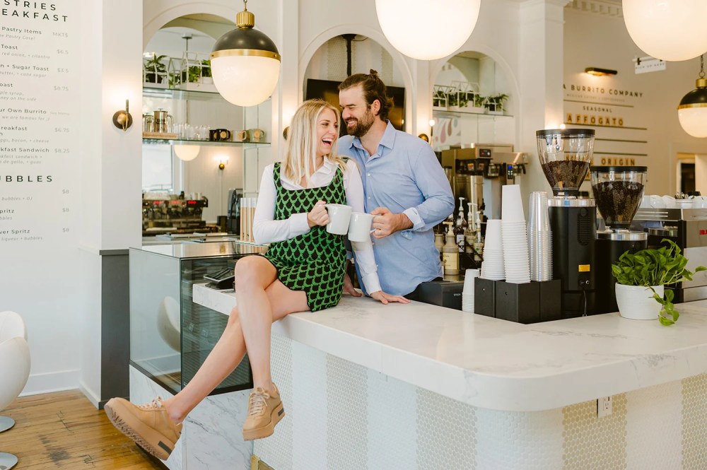 Man and woman laughing in a cafe, sitting on a countertop with mugs.