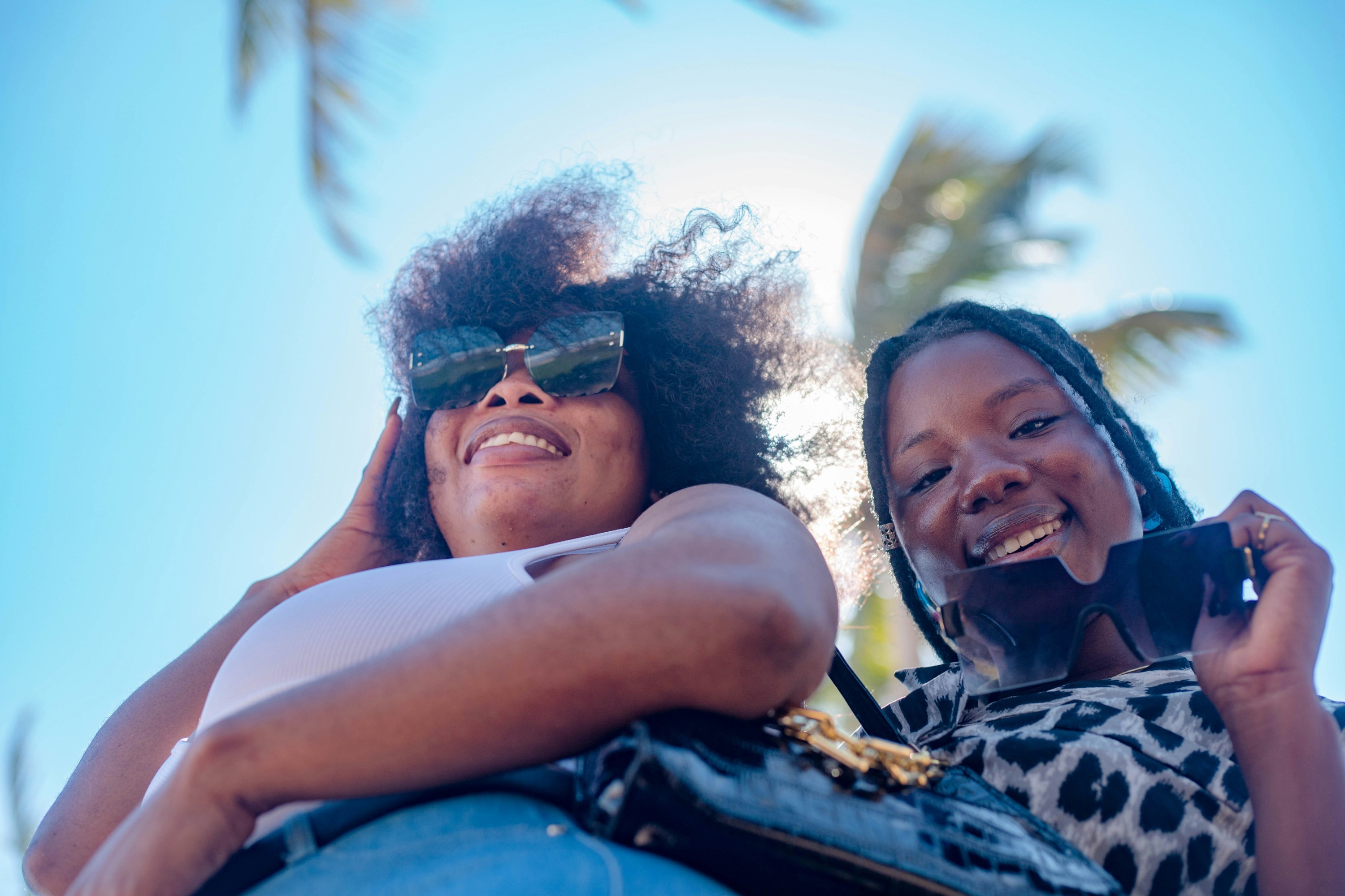 Two people smiling up at the camera, holding sunglasses, with a palm tree and blue sky in the background.