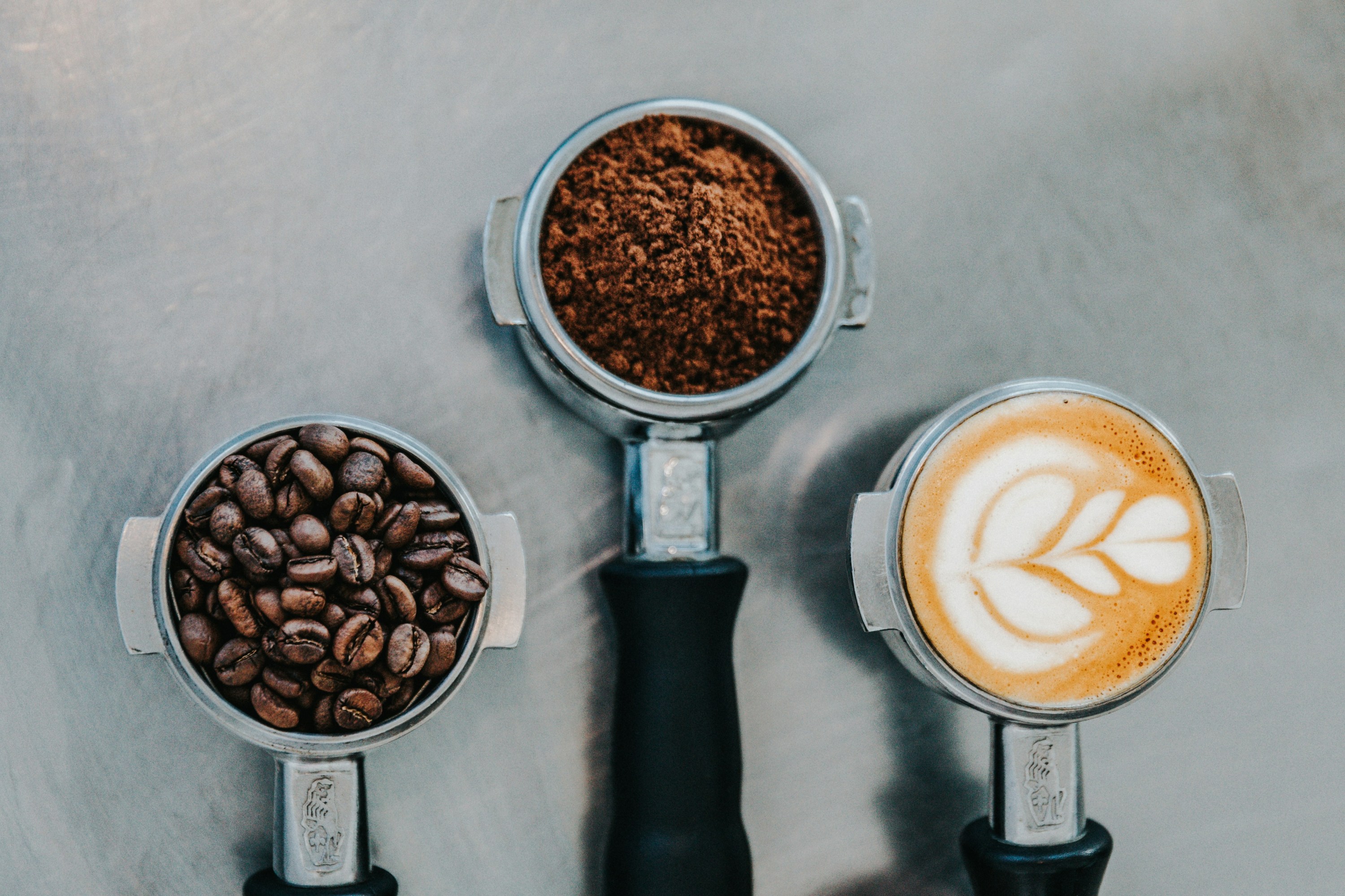 Three portafilters with coffee beans, ground coffee, and latte art on a table.