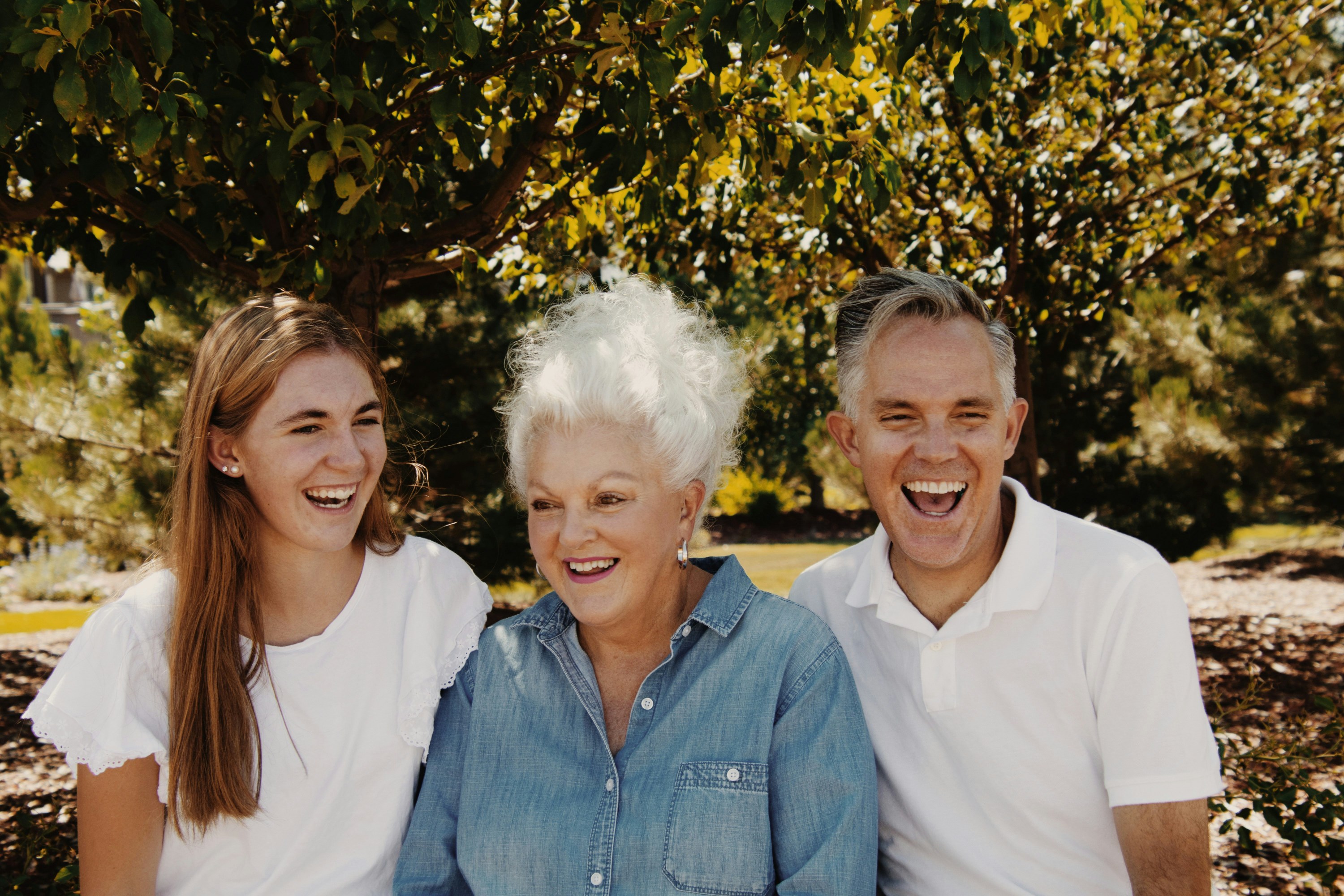 Three people laughing under a tree, wearing casual white and blue clothing.