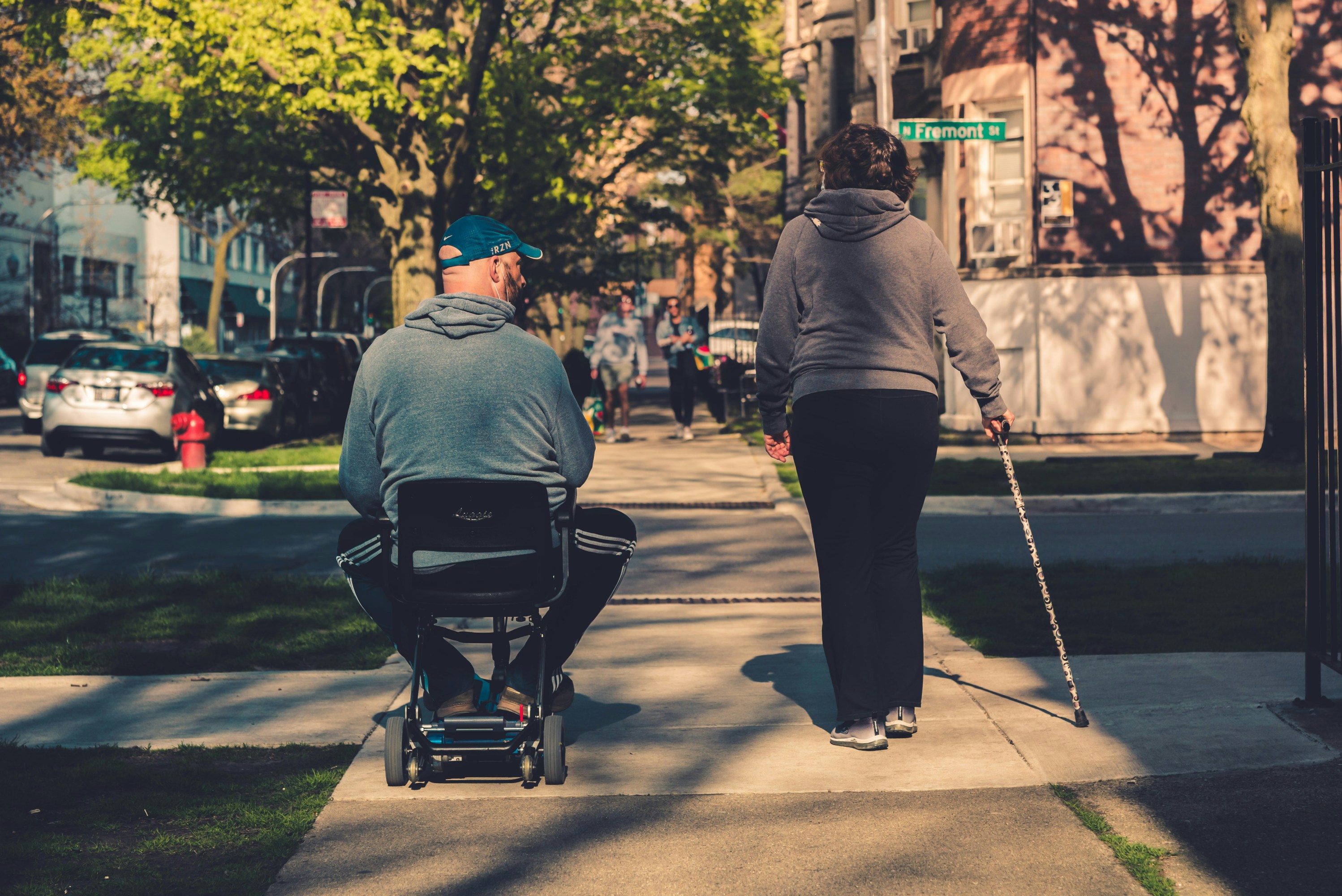 Two people on a sidewalk, one sitting on a mobility scooter, the other walking with a cane.